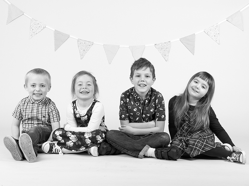 A group of four children sitting down in a line all looking at the camera smiling with bunting behind them.