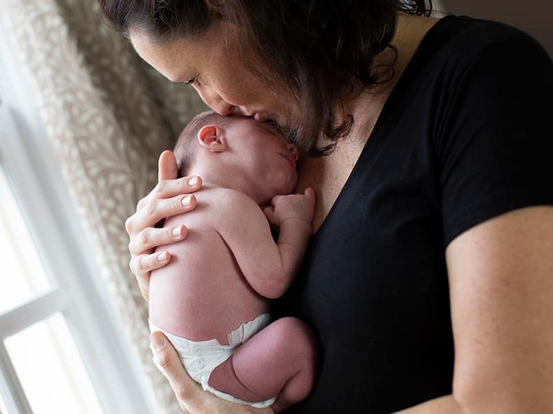 Portrait of a mother holding her newborn baby close to her chest close to a window.