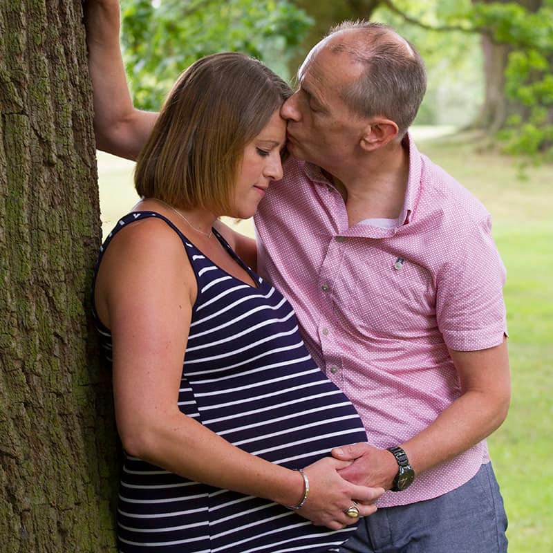 Close up shot of couple leaning against tree both touching her baby bump