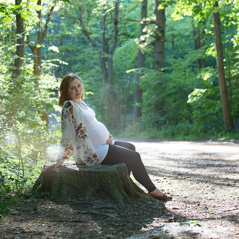 Portrait of a pregnant woman sitting on a log in a wood full of sunlight.