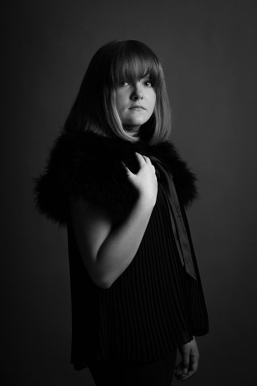 Black and white portrait of teen wearing black standing to the left holding her right hand up to her right shoulder.