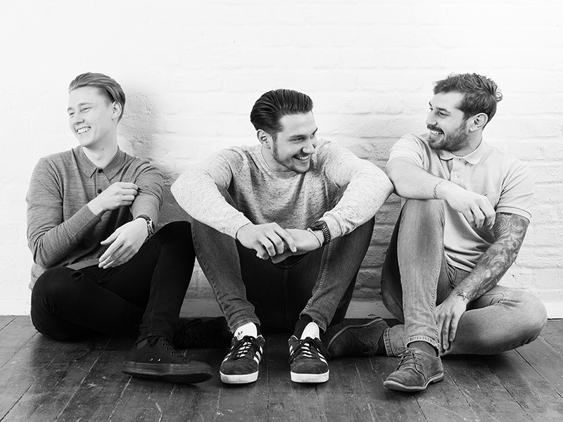 Three brothers sitting down laughing and leaning against a white brick wall at the studio.