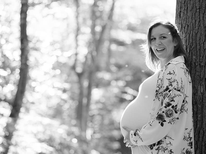 Pregnant woman leaning against a tree smiling in the sunlight.